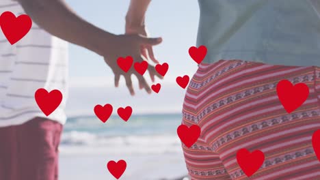 Animation-of-red-hearts-over-midsection-of-african-american-couple-holding-hands-on-beach