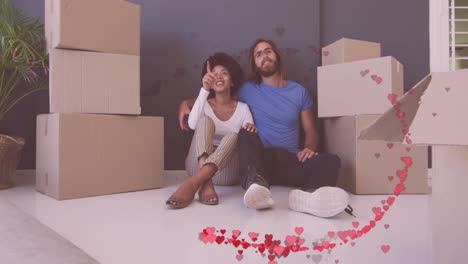 Animation-of-red-hearts-over-happy-diverse-couple-sitting-amongst-boxes-after-moving-into-house