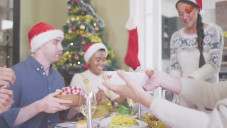 Animation-of-hearts-over-diverse-friends-sitting-at-table-at-christmas