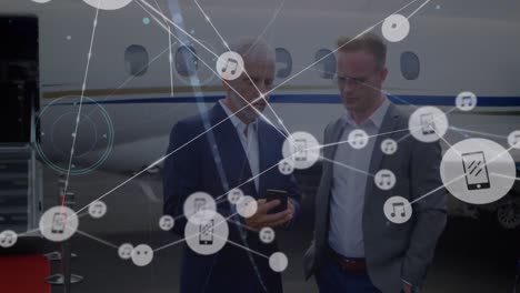 Animation-of-network-of-digital-icons-over-two-caucasian-businessmen-discussing-at-airport