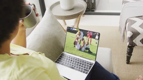 Video-of-biracial-woman-sitting-on-sofa-and-watching-football-on-laptop-at-home