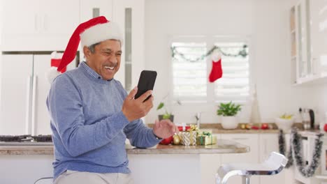 Video-of-happy-senior-biracial-man-in-santa-hat-making-christmas-video-call-on-smartphone,-waving