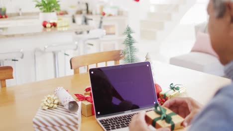 Video-of-senior-biracial-man-holding-gift-making-laptop-christmas-video-call,-copy-space-on-screen