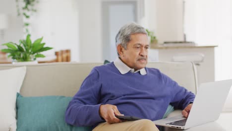 Video-Eines-älteren-Gemischtrassigen-Mannes,-Der-Smartphone-Und-Laptop-Benutzt-Und-Lächelnd-Im-Wohnzimmer-Sitzt