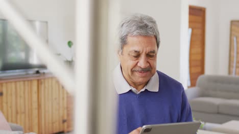 Video-Eines-älteren-Gemischtrassigen-Mannes,-Der-Lächelnd-Im-Wohnzimmer-Sitzt-Und-Ein-Tablet-Benutzt