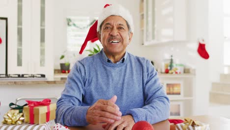 Video-of-happy-senior-biracial-man-in-santa-hat-making-christmas-video-call,-waving-to-camera