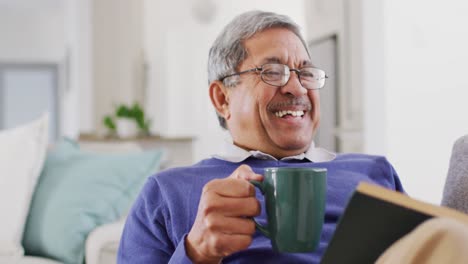 Video-of-happy-senior-biracial-man-drinking-coffee,-reading-book-at-home-and-laughing