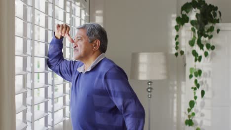 Video-of-thoughtful-senior-biracial-man-standing,-leaning-and-looking-out-of-window