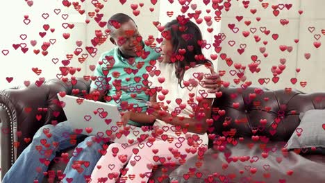 Animation-of-red-heart-icons-floating-over-african-american-couple-using-laptop-on-the-couch-at-home