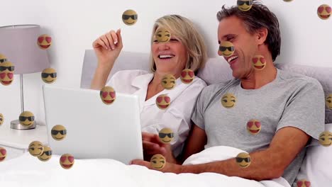 Animation-Von-Gesichts-Emoji-Symbolen-Vor-Einem-Kaukasischen-älteren-Paar,-Das-Auf-Einem-Laptop-Im-Bett-Einen-Videoanruf-Führt