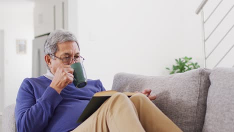 Video-Eines-Glücklichen-älteren-Birazialen-Mannes,-Der-Zu-Hause-Auf-Der-Couch-Kaffee-Trinkt-Und-Ein-Buch-Liest