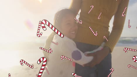 Animation-of-candy-canes-over-biracial-woman-with-her-daughter-at-beach