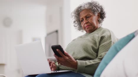 Portrait-of-senior-african-american-woman-sitting-on-sofa,-using-laptop-and-smartphone