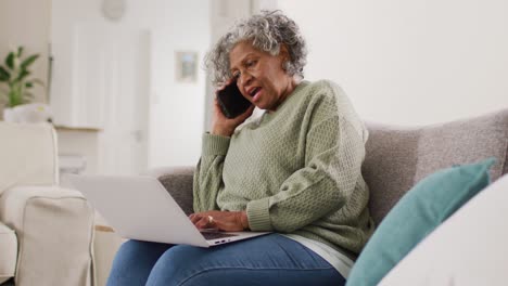 Portrait-of-senior-african-american-woman-sitting-on-sofa,-using-laptop-and-talking-on-smartphone