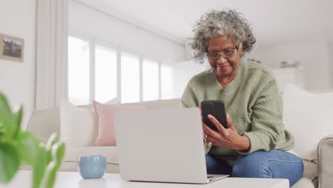 Portrait-of-happy-senior-african-american-woman-sitting-on-sofa,-using-laptop-and-smartphone