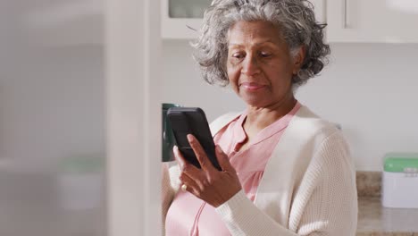 Porträt-Einer-Glücklichen-älteren-Afroamerikanischen-Frau,-Die-Ihr-Smartphone-Benutzt-Und-In-Der-Küche-Kaffee-Trinkt