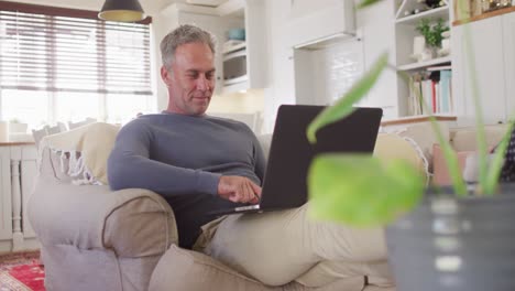 Happy-caucasian-man-sitting-on-sofa-in-living-room,-using-laptop