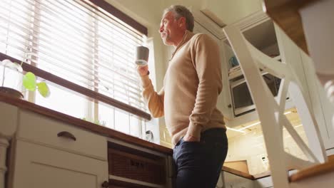 Happy-caucasian-man-looking-through-window-and-drinking-coffee-in-kitchen