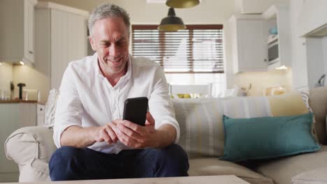 Happy-caucasian-man-sitting-on-sofa-in-living-room,-using-smartphone