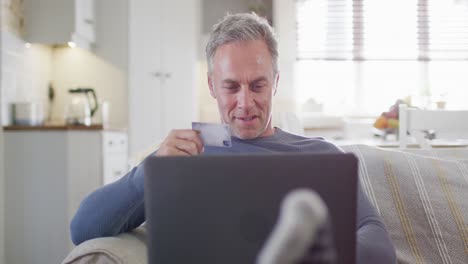 Happy-caucasian-man-sitting-on-sofa-in-living-room,-using-laptop-and-holding-credit-card