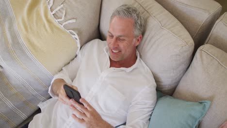 Happy-caucasian-man-lying-on-sofa-in-living-room,-using-smartphone