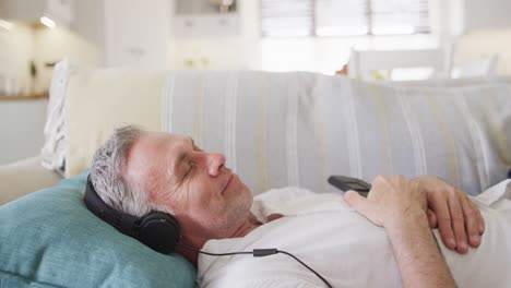 Happy-caucasian-man-lying-on-sofa-in-living-room,-using-smartphone-and-headphones