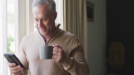 Happy-caucasian-man-using-smartphone-and-drinking-coffee-in-bedroom