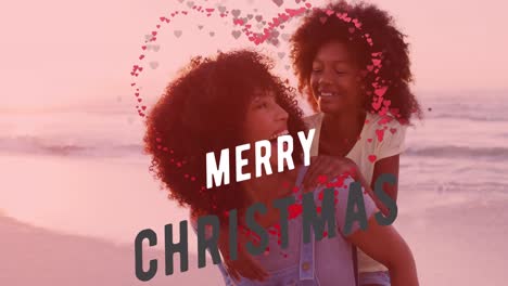 Animation-of-merry-christmas-text-over-happy-african-american-mother-and-daughter-on-sunny-beach