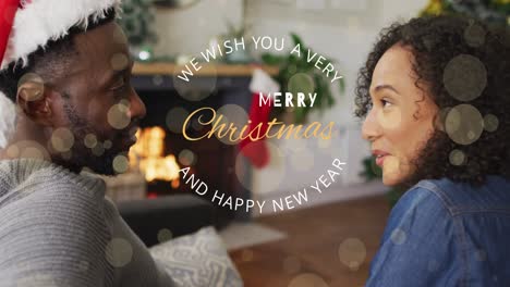 Animation-of-merry-christmas-and-new-year-text-over-happy-diverse-couple-talking-by-fireplace