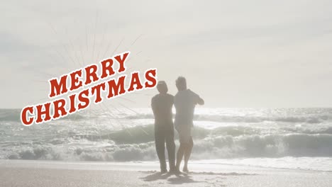 Animation-of-merry-christmas-text-in-red-over-happy-senior-biracial-couple-dancing-on-sunny-beach