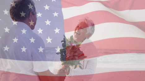 Animation-of-flag-of-united-states-of-america-over-senior-biracial-couple-with-flowers-on-beach