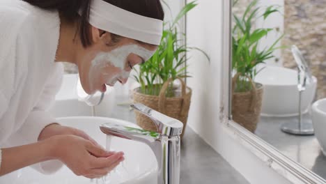 Biracial-woman-with-mask-washing-face-in-bathroom