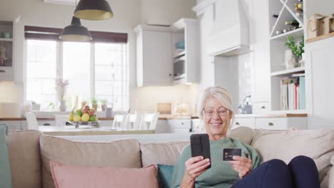 Glückliche-ältere-Kaukasische-Frau,-Die-Auf-Dem-Sofa-Sitzt,-Ihr-Smartphone-Benutzt-Und-Eine-Kreditkarte-In-Der-Hand-Hält