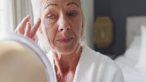 Happy-senior-caucasian-woman-on-bed-in-bedroom,-looking-at-mirror,-applying-cream-on-her-face