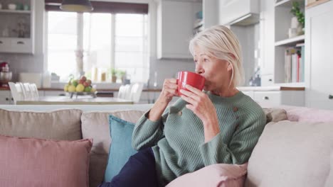 Thoughtful-senior-caucasian-woman-sitting-on-sofa-in-living-room,-drinking-coffee