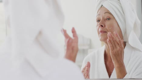 Happy-senior-caucasian-woman-looking-at-mirror-in-bathroom-and-applying-cream-on-her-face
