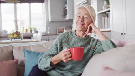 Portrait-of-happy-senior-caucasian-woman-sitting-on-sofa-in-living-room,-drinking-coffee