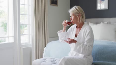 Happy-senior-caucasian-woman-sitting-on-bed-in-bedroom,-drinking-tea