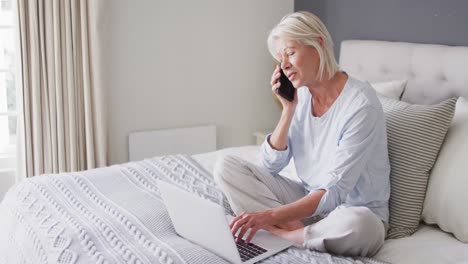 Happy-senior-caucasian-woman-sitting-on-bed-in-bedroom,-using-laptop-and-talking-on-smartphone