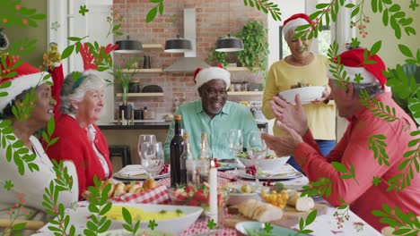 Animation-of-christmas-holly-over-senior-diverse-friends-in-santa-hats-at-christmas-meal-table