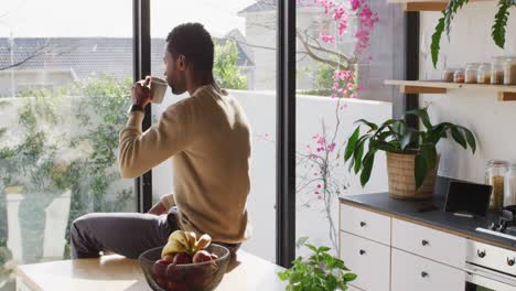 Happy-african-american-man-sitting-at-countertop-in-kitchen,-drinking-coffee