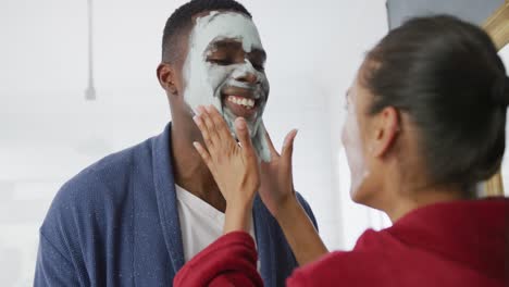 Video-of-happy-diverse-couple-having-fun-applying-cleansing-face-masks-and-laughing-in-bathroom