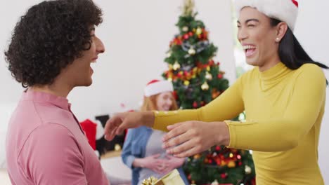 Happy-biracial-man-receiving-present-and-hugging-non-binary-transgender-friend-in-santa-hat-at-home