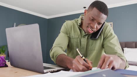 Video-Eines-Afroamerikanischen-Mannes,-Der-Laptop-Benutzt,-Smartphone-Spricht-Und-Notizen-Schreibt-Und-Im-Schlafzimmer-Arbeitet