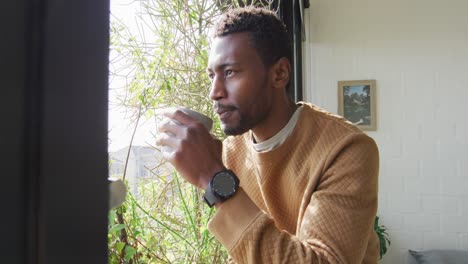 Happy-african-american-man-looking-through-window-and-drinking-coffee