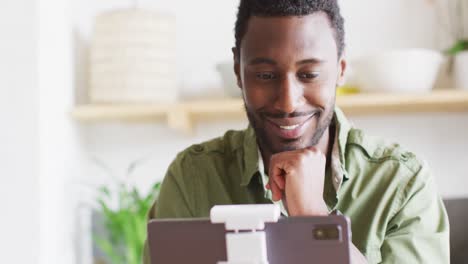 Happy-african-american-man-sitting-at-table-in-kitchen,-using-tablet-for-video-call