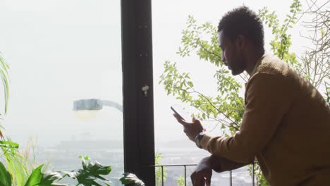 Glücklicher-Afroamerikanischer-Mann,-Der-Durch-Das-Fenster-Schaut-Und-Sein-Smartphone-Benutzt