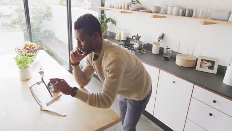 Happy-african-american-man-leaning-on-countertop-in-kitchen,-using-tablet-and-talking-on-smartphone