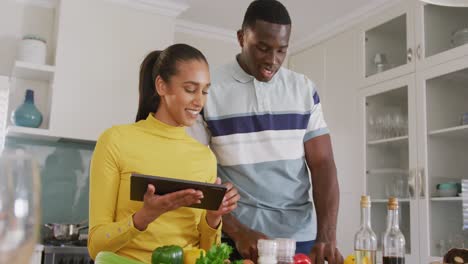 Video-of-happy-diverse-couple-using-recipe-on-tablet-preparing-food-together-in-kitchen