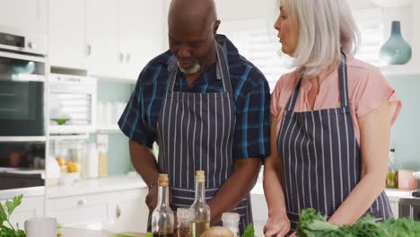 Happy-senior-diverse-couple-wearing-aprons-and-cooking-in-kitchen